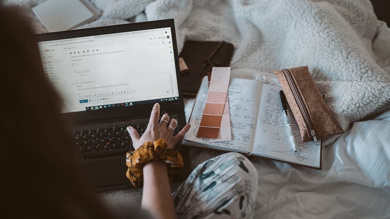 An over-the-shoulder view of a person working at their laptop on their bed.